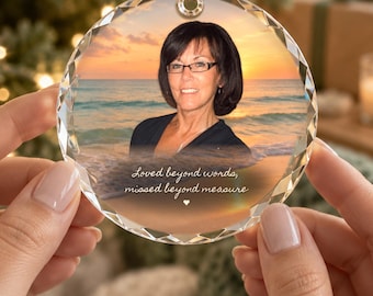 May include: A circular crystal ornament with a faceted edge featuring a photograph of a person with dark hair and glasses against a sunset beach background. The text reads, Loved beyond words, missed beyond measure, with a small heart icon.