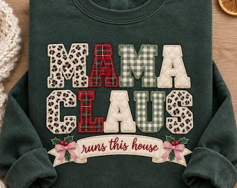 May include: A dark green crewneck sweatshirt featuring the text MAMA CLAUS in large, multi-patterned fabric letters. Below the text is a banner with the script words runs this house, decorated with small red berries and green leaves.