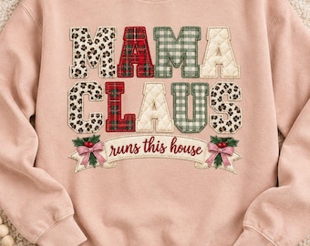 May include: A light pink crewneck sweatshirt featuring the text MAMA CLAUS in large, multi-patterned, stitched-on letters. Below the text, a banner reads runs this house, flanked by embroidered holly leaves, red berries, and pink bows.