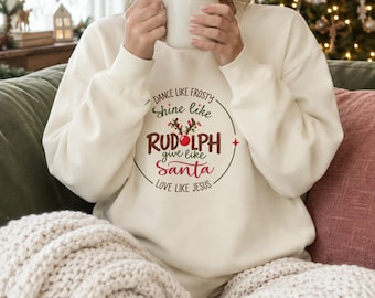 May include: A cream-colored crewneck sweatshirt featuring the text DANCE LIKE FROSTY, SHINE LIKE RUDOLPH, give like Santa, LOVE LIKE JESUS, with a graphic of reindeer antlers and a red nose centered within a circular design.