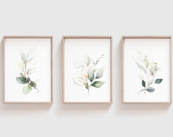 Puede incluir: Tres impresiones botánicas enmarcadas con ilustraciones en acuarela de hojas y follaje en tonos verdes, dorados y blancos. La obra de arte está sobre un fondo blanco, cada impresión está dentro de un marco de madera marrón claro.