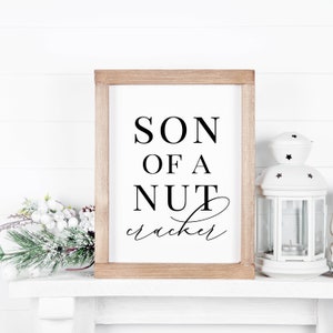 May include: A wooden sign with the text "SON OF A NUTcracker" in black lettering on a white background.
