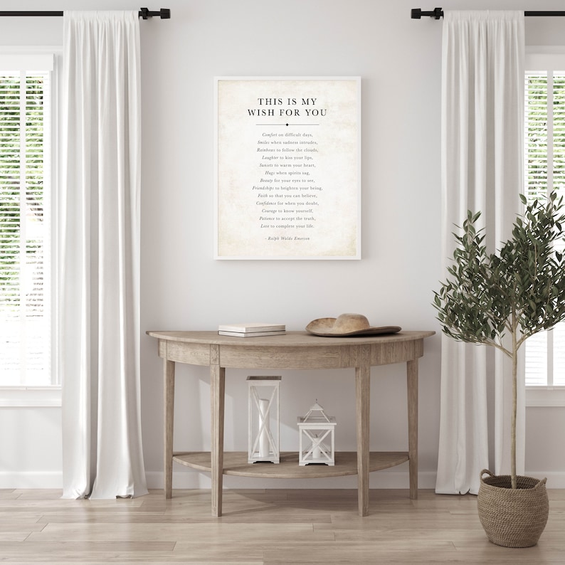May include: A white framed print with a black and white inspirational quote that reads "This is my wish for you." The quote is by Ralph Waldo Emerson. The print is hanging on a white wall above a wooden console table with two white lanterns.