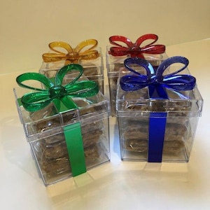 May include: Four clear plastic gift boxes, each filled with cookies. The boxes are adorned with colorful bows in yellow, red, green, and blue. Each box has a matching ribbon. The cookies are stacked inside the boxes.