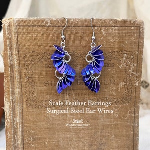 May include: A pair of scale feather earrings with surgical steel ear wires. The earrings feature iridescent blue scales arranged in a cascading design. The earrings are displayed on a vintage book.