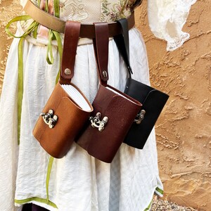 May include: Three leather pouches in brown, dark brown, and black, each with a metal clasp, hang from a belt. The pouches are attached to the belt with leather straps. The background is a textured, light brown wall.