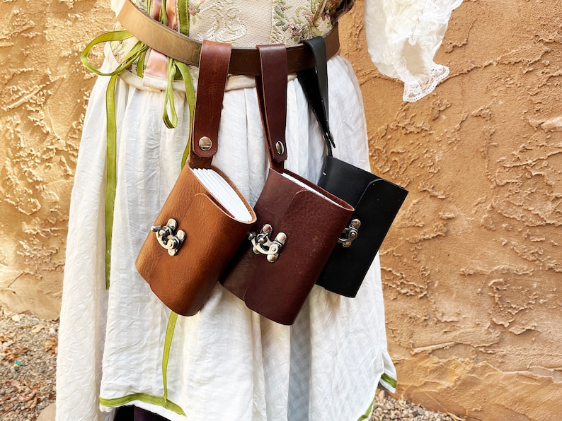 May include: Three leather pouches in brown, dark brown, and black, each with a metal clasp, hang from a belt. The pouches are attached to the belt with leather straps. The background is a textured, light brown wall.