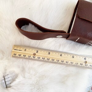 May include: A brown leather case with a handle and silver hardware, approximately 6 inches long, rests on a white, textured surface. A wooden ruler, marked in inches, is placed next to the case.