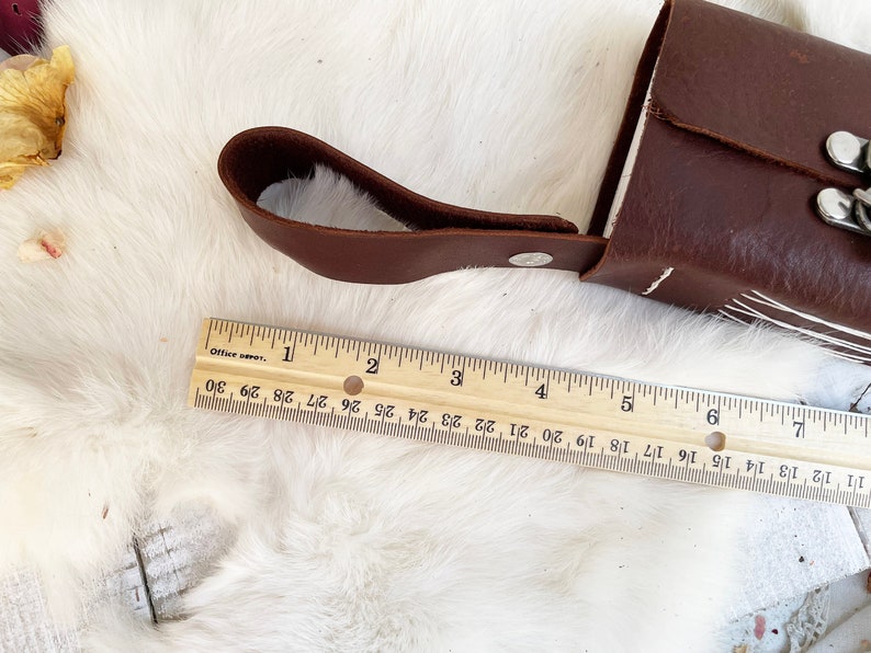May include: A brown leather case with a handle and silver hardware, approximately 6 inches long, rests on a white, textured surface. A wooden ruler, marked in inches, is placed next to the case.
