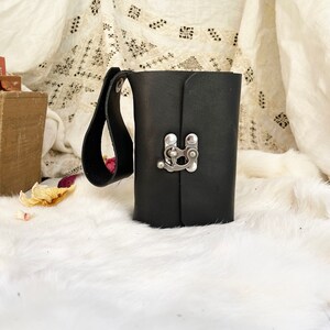 May include: A black leather journal with a silver clasp and a handle. The journal is rectangular and closed, with a strap attached to the side. The background includes books and a white, fluffy surface.