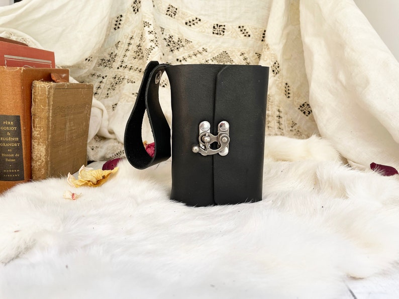 May include: A black leather journal with a silver clasp and a handle. The journal is rectangular and closed, with a strap attached to the side. The background includes books and a white, fluffy surface.