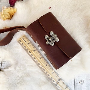 May include: A small, rectangular, dark brown leather journal with a silver clasp and a matching leather strap. The journal is approximately 4 inches long. The journal is closed and resting on a white, fluffy surface.