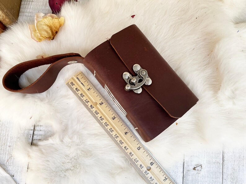 May include: A small, rectangular, dark brown leather journal with a silver clasp and a matching leather strap. The journal is approximately 4 inches long. The journal is closed and resting on a white, fluffy surface.