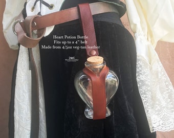 Puede incluir: Una botella de poción en forma de corazón con un tapón de corcho, sujeta por un arnés de cuero marrón sujeto a un cinturón. El texto de la imagen dice: "Heart Potion Bottle Fits up to a 4" belt Made from 4/5oz veg-tan leather."