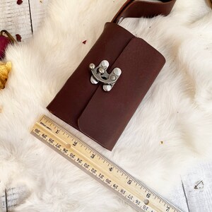 May include: A dark brown leather journal with a silver clasp and a leather strap. The journal is approximately 6 inches long. The journal is on a white fur-like surface with a wooden ruler.