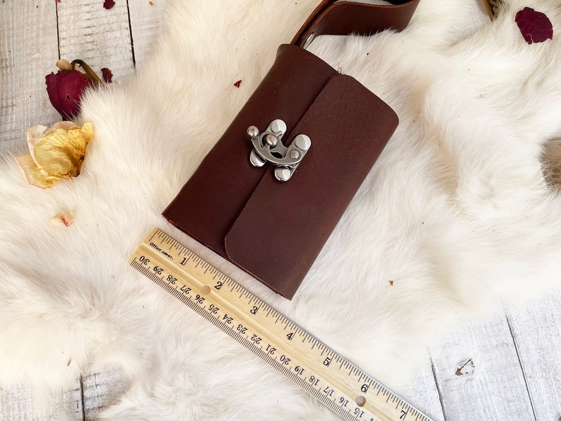 May include: A dark brown leather journal with a silver clasp and a leather strap. The journal is approximately 6 inches long. The journal is on a white fur-like surface with a wooden ruler.