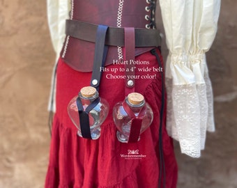 Puede incluir: Dos botellas de poción de vidrio en forma de corazón con tapones de corcho y cintas, suspendidas de un cinturón. Una botella tiene una cinta negra, la otra una cinta burdeos. El texto "Heart Potions" y "Se adapta a cinturones de hasta 10 cm de ancho" es visible.