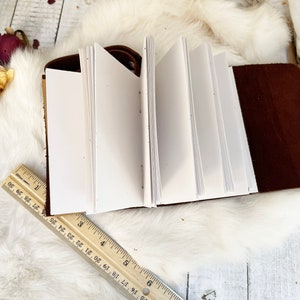 May include: Open leather journal with blank white pages. The journal is brown with a snap closure. A wooden ruler is placed near the journal. The journal is on a white fluffy surface with dried flowers.