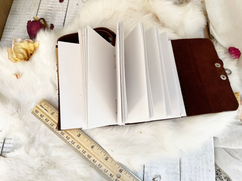 May include: Open leather journal with blank white pages. The journal is brown with a snap closure. A wooden ruler is placed near the journal. The journal is on a white fluffy surface with dried flowers.