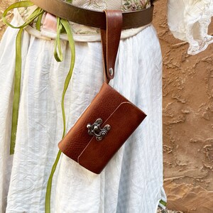 May include: A brown leather pouch with a metal clasp, attached to a brown leather belt. The pouch is rectangular and has a textured surface. The belt is worn over a white dress with green ribbons.