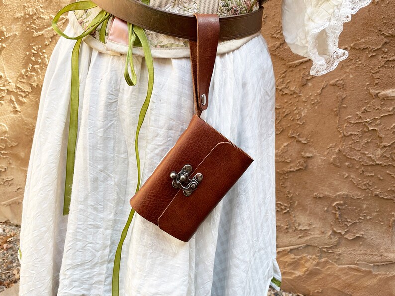 May include: A brown leather pouch with a metal clasp, attached to a brown leather belt. The pouch is rectangular and has a textured surface. The belt is worn over a white dress with green ribbons.