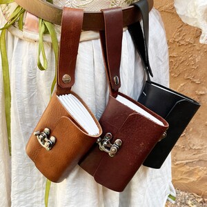 May include: Three leather pouches in varying shades of brown and black, each with a small notebook inside. The pouches are attached to a brown belt with a strap and metal clasp closure. The notebooks are visible inside the pouches.