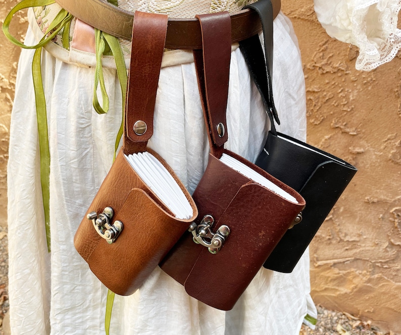 May include: Three leather pouches in varying shades of brown and black, each with a small notebook inside. The pouches are attached to a brown belt with a strap and metal clasp closure. The notebooks are visible inside the pouches.