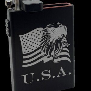 May include: Black metal container with a hinged lid and a built-in grinder. The container features an engraved image of an American bald eagle with a flag and the text "U.S.A."