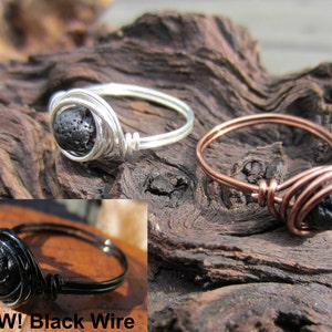 May include: Three rings with black lava stone beads. One ring is silver wire, another is copper wire, and the third is black wire. The rings are displayed on a piece of wood. The text "NEW! Black Wire" is visible.