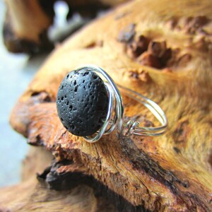 Puede incluir: Anillo artesanal con una piedra de lava negra porosa engastada en un alambre plateado. El diseño del anillo muestra la textura natural de la piedra, creando una estética única y terrosa. El anillo se exhibe sobre un trozo de madera.