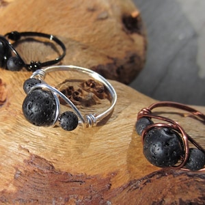 May include: Three rings with black lava rock beads. One ring is black, one is silver, and one is copper. The rings are displayed on a piece of wood.