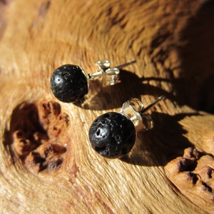 May include: A pair of black lava rock stud earrings with silver posts. The earrings are on a wooden surface.