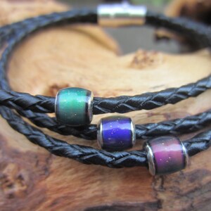 May include: Braided black leather bracelet with three color-changing beads. The beads shift from green to purple and blue, each with a silver-colored metal end. The bracelet is wrapped around itself, creating a layered effect.