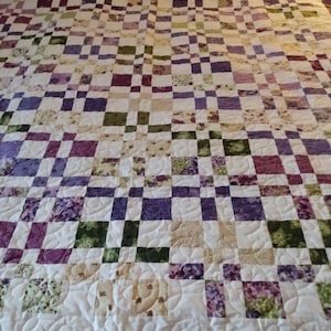 May include: A purple, green, and cream quilt with a floral pattern. The quilt is made with squares and has a quilted design.