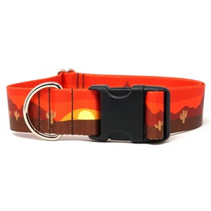 May include: Orange dog collar with a desert sunset design. The collar features a black plastic buckle, a silver metal D-ring, and a repeating pattern of cacti and mountains. The collar is approximately 1 inch wide.