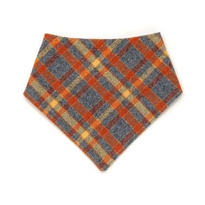 May include: A triangular pet bandana featuring a plaid pattern with intersecting lines in shades of burnt orange, slate blue, and muted yellow. The fabric has a textured appearance, and the accessory is shaped to drape into a point at the bottom.