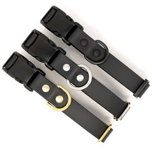 May include: Three black dog collars with different metal hardware. One collar has a black metal D-ring, one has a silver metal D-ring, and the third has a gold metal D-ring. Each collar has a black plastic buckle and is made of black leather.