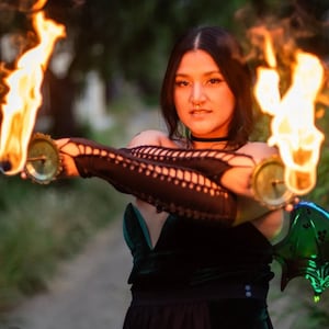 May include: A person wearing a black and green outfit with intricate lace sleeves is performing a fire dance. They are holding two flaming torches in each hand. The torches are attached to a metal base with a circular design.