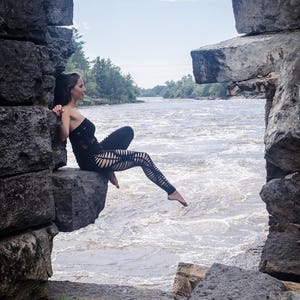 以下が含まれることがあります： 黒いクロシェの衣装を着た女性が、激流の川を見下ろす岩の崖に座っています。川は木々に囲まれ、空は曇っています。