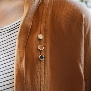 Può includere: Primo piano di una giacca marrone con tre bottoni rotondi decorativi nei toni del beige, marrone e nero. La giacca è indossata sopra una camicia a righe bianche e blu navy.