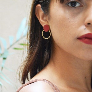 Può includere: Una donna indossa un paio di orecchini a cerchio dorati con un ciondolo rotondo rosso. Gli orecchini sono semplici ed eleganti.