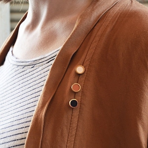 Può includere: Un cardigan marrone con una fila di tre piccoli spilli rotondi. Gli spilli sono di diversi colori: bianco, arancione e nero.