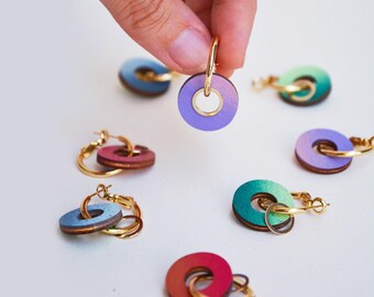 Puede incluir: Una colección de pendientes de aro de madera coloridos. Los pendientes presentan un diseño circular de madera con un degradado de colores, incluyendo morado, azul, verde y rojo, combinados con aros dorados. Los pendientes se muestran sobre una superficie blanca.
