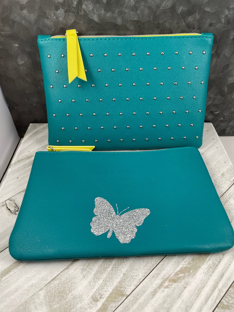 May include: Two teal cosmetic bags. The top bag has silver studs and a yellow zipper pull. The bottom bag has a silver glitter butterfly design. Both bags have yellow zippers.