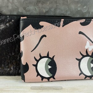 May include: Two cosmetic bags. One is black and covered in sequins. The other is pink with a cartoon face design, black accents, and a white butterfly. Both have black zippers and the text "Jenns Perfect Accessories".