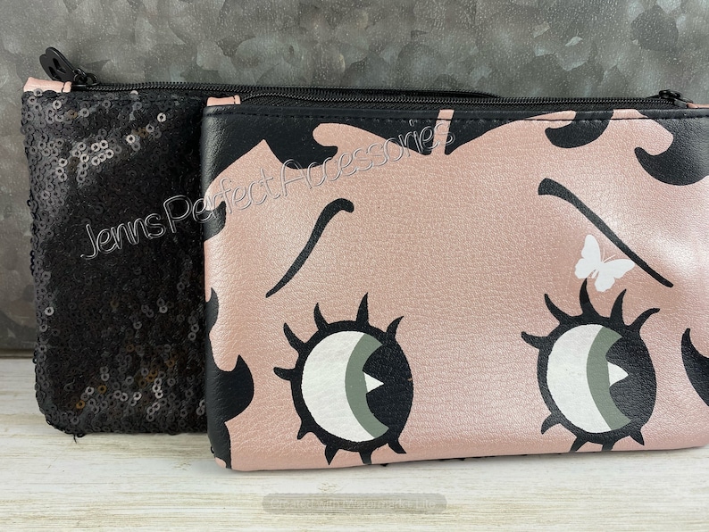 May include: Two cosmetic bags. One is black and covered in sequins. The other is pink with a cartoon face design, black accents, and a white butterfly. Both have black zippers and the text "Jenns Perfect Accessories".