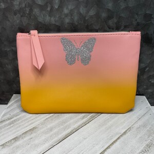 May include: A pink and yellow ombre cosmetic bag with a silver glitter butterfly design. The bag has a zipper closure and a pink pull tab. The bag is sitting on a white wooden surface.