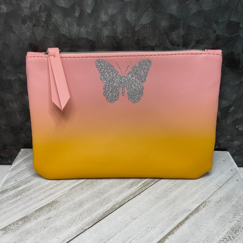 May include: A pink and yellow ombre cosmetic bag with a silver glitter butterfly design. The bag has a zipper closure and a pink pull tab. The bag is sitting on a white wooden surface.