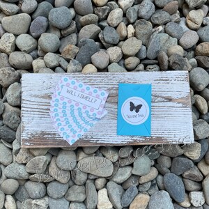 May include: A rustic wooden sign with white paint and a distressed look, resting on a bed of stones. The sign holds a stack of cards with the text "WILL I SMELL?" and a small blue box with a butterfly logo and the text "Tips and Trick."