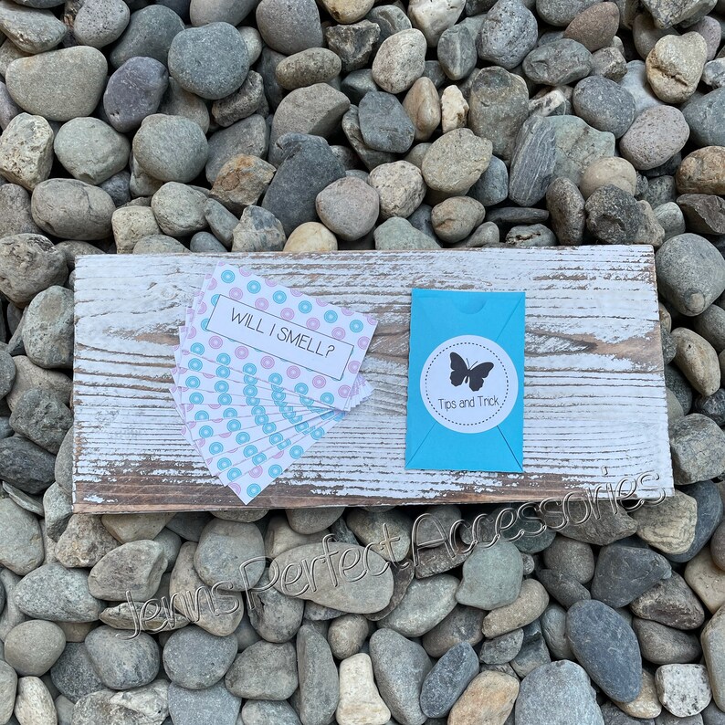 May include: A rustic wooden sign with white paint and a distressed look, resting on a bed of stones. The sign holds a stack of cards with the text "WILL I SMELL?" and a small blue box with a butterfly logo and the text "Tips and Trick."